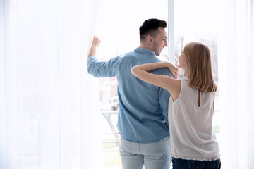 Happy couple standing near window and opening curtains