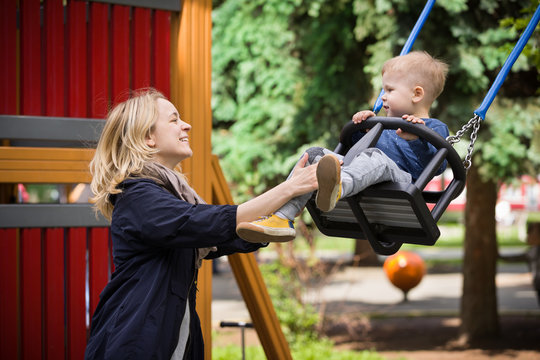 Happy Mother Pushing Cute Toddler Son On Swing In A Park. Mom And Her Child Playing Together In Nature Outdoors. Happy Loving Family Having Fun.