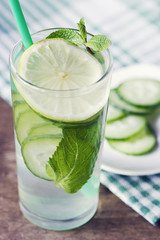 A glass with straws with detox water from cucumber, mint and lemon stands on a wooden background with a checkered green towel