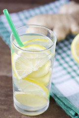 A glass with straws with detox water from the root of ginger and lemon stands on a wooden background with a checkered green towel