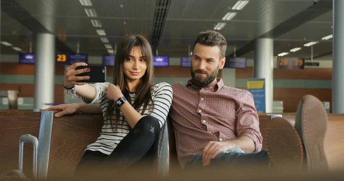 Happy young couple in love using smart phone for taking selfie while sitting at airport lounge.