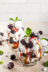 Traditional English dessert. Eton mess - whipped cream, meringue, fresh blackberries, sauce and caramel. In serving glasses on a light stone table. Copy space