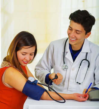 Close Up Of A Handsome Smiling Young Doctor With Stethoscope Around His Neck Taking The Pulse To A Young Woman With A Tensiometer, In A Doctor Consulting Room Background