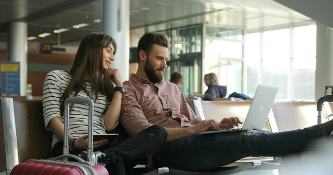 Happy young beatiful loving couple is sitting at airport lounge. Woman using tablet while man using laptop.