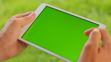 Male hands holding white tablet computer device with green screen on green grass background. Outdoors in park. Man scrolling, zooming, tapping on touchscreen. Chroma key. Close up.