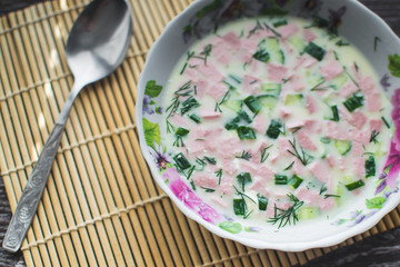okroshka in a bowl near the window on a black wooden background, lying next to a spoon