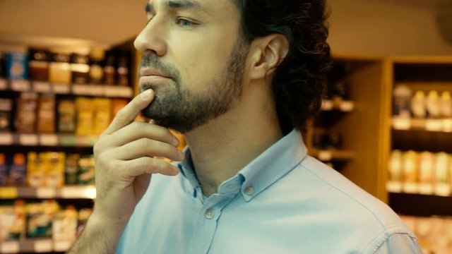 Portrait of young caucasian man with curly hair trying to choose good bottle of wine in the supermarket. Close up.