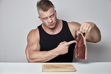 Nahaufnahme großer Bodybuilder isst großes Rindersteak hinter dem Tisch gegenüber der grauen Wand