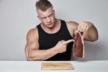 Deurstickers Steakhouse Close-up grote bodybuilder eet grote biefstuk achter tafel tegenover grijze muur
