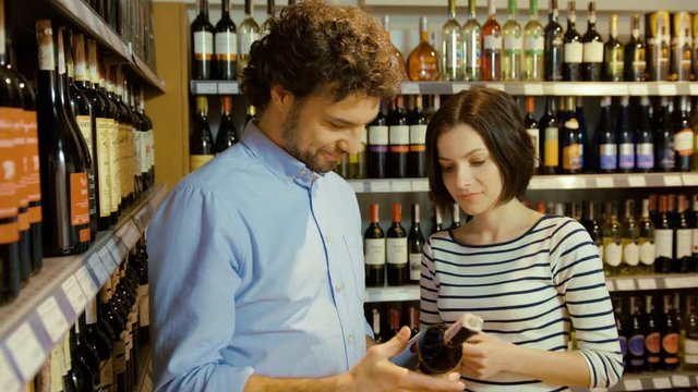 Attractive couple trying to choose bottle of wine in the supermarket. Man taking one bottle, than anouther, young woman choosing one of this bottle.