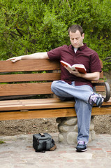 Man sitting on a bench reading a book with his bag on the ground.