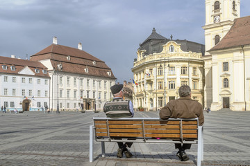 Obraz premium Two people sitting on a bench in the famous Piata Mare, Large Square, in Sibiu, Romania, rest and observe passersby