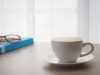 wood office table with cup of latte coffee and modern eyeglasses on blurry white curtain texture background.