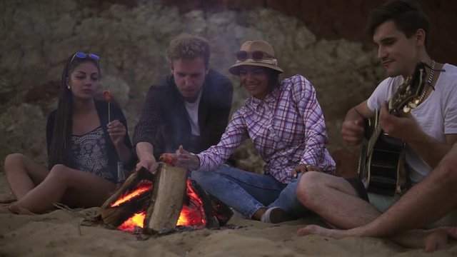 Group of young and cheerful people sitting by the fire on the beach in the evening, singing songs and playing guitar. Picninc on the beach in the evening. Slowmotion shot