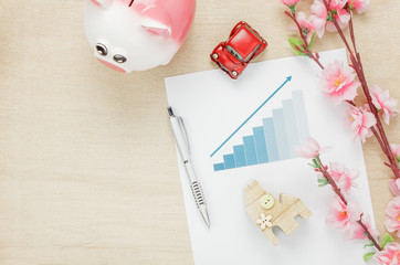 Top view business office desk concept. Saving money with charted summary  Wood house  also car and clock on wooden shelf.The beautiful pink flower with pen on wood background with copy space.