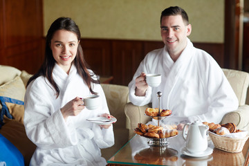 Young couple in bathrobes having coffee with buns in hotel room