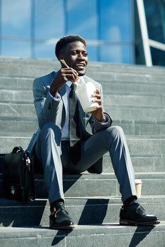 Happy businessman with chopsticks eating food brom box on staircase