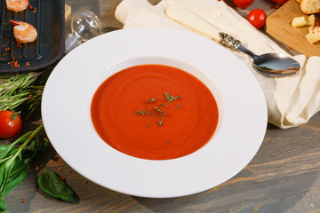 Red tomato soup gazpacho in a large white plate on a table decorated with ingredients. Leaves of basil and rosemary, fried prawns on a black frying pan, cherry tomatoes, croutons and parmesan cheese.