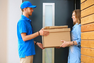Smiling delivery man in blue uniform delivering parcel box to recipient - courier service concept - obrazy, fototapety, plakaty