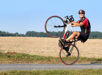 Obraz premium Cheerful cyclist riding on the rear wheel. Biker balancing while driving on a road bike. Risky ride on one wheel.