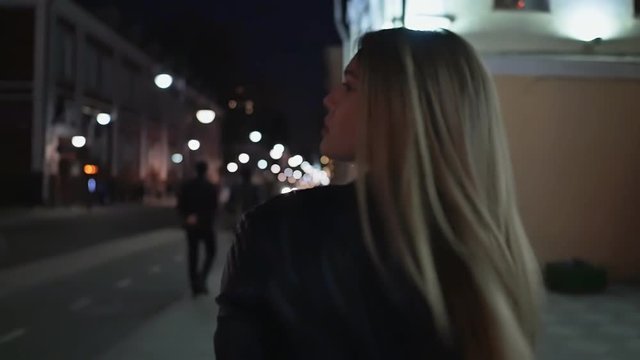 Beautiful young woman against night shined city. The blonde is walking around the night city. Shooting from behind.