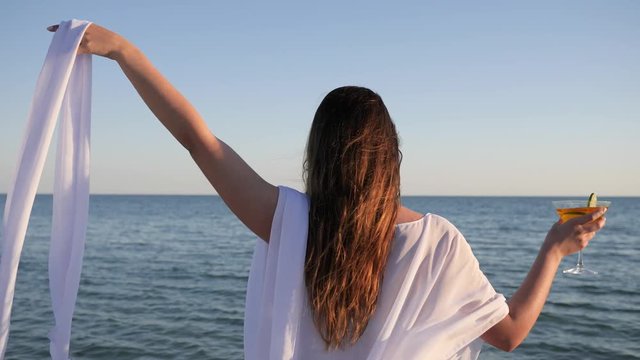 girl in Swimsuit on shore, women on background water Waving white cloth, holiday near ocean, female drinking Colored cocktail with fruit in Coast, weekend on Tropical islands, summer rest