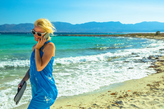 Happy woman talking on a smartphone walking on the beach. Beach woman wearing an blue sarong, sunglasses and holding sea slippers enjoying. Turquoise sea of famous Elafonisos island, Greece, behind.