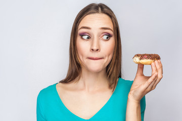 Portrait of beautiful girl with chocolate donuts. thoughtful and looking at donut with big confused eyes. studio shot on light gray background.