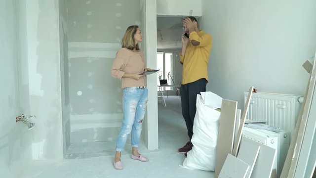 Sad, unhappy couple talking on cellphone standing in room at their new home
