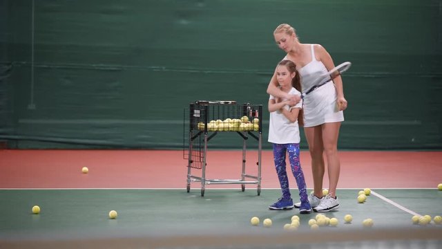 The young coach teaches to keep the child's racket correctly, the girl actively waves her hand to learn how to beat off a tennis ball