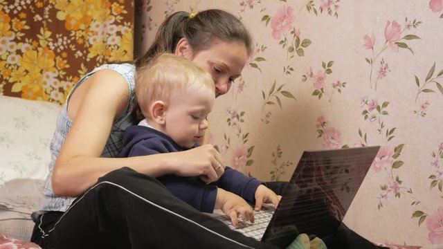 Young woman with her son sitting at home on the couch with a laptop. Look carefully at the screen and press the keys.