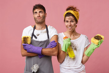 Husband wearing apron and gloves holding sponge looking fatigued up after helping his wife to clean house. Housewife with dirty face and clothes having indignant look standing near her lover