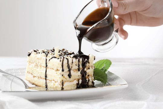 Female hand pouring chocolate sauce onto plate with tasty cake on table