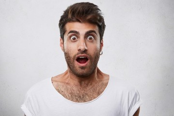 Portrait of stylish bearded guy having trendy hairstyle wearing earring and white T-shirt looking with his eyes popped out and opened mouth having shock and scared look. People and emotions concept
