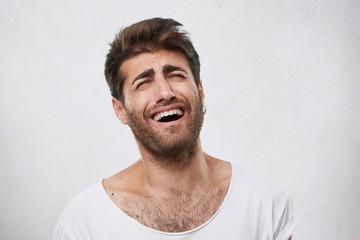 Bearded man with thick eyebrows, fashionable hairstyle and beard bursting into laughing after hearing funny joke. Attractive man closing his eyes with laughing. People and facial expressions concept