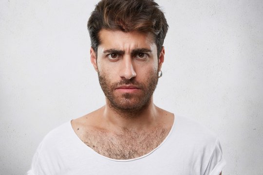 Close up portrait of serious gloomy bearded man with stylish hairstyle posing against white background. Handsome guy with dark eyes frowning his face showing his disatisfaction and dipleasment