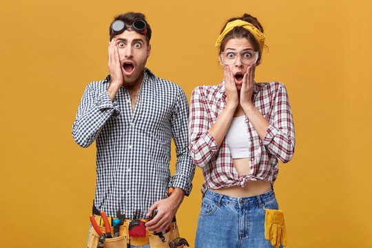 Renovation, repair, maintenance and remodeling. Studio shot of shocked repairman with belt kit of tools screaming in astonishment, standing next to female colleague, both wearing protective goggles