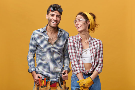 Cheerful female electrician or mechanic in protective gloves and goggles looking at her male colleague who is wearing belt kit with pliers, screwdriver, hammer and other tools needed for work