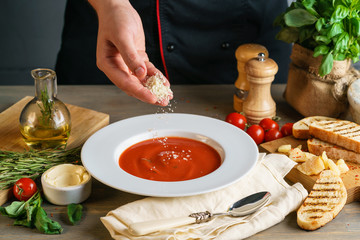 The cook sprinkles tomato cream soup with chopped parmesan cheese. The kitchen table is decorated with ingredients.