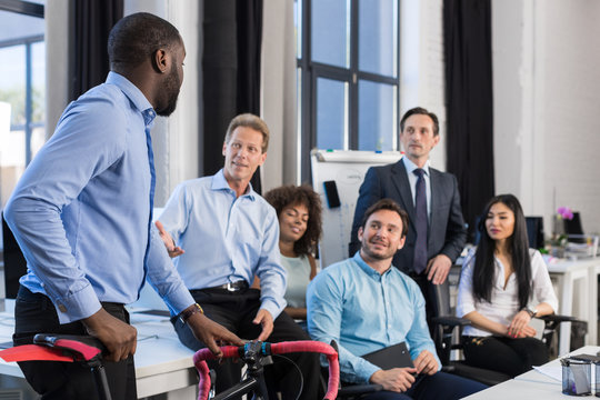 Business People Group In Creative Office, African American Businessman Hold Bicycle, Team Meeting And Communicating Sitting At Desk In Modern Coworking Space, Mix Race Businesspeople Working Together