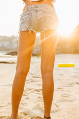 Close up of legs of a beautiful slim lady in white shirt and denim shorts walk to big inflatable ring. She hold hands on her back. Beauty sunshine cute girl on a tropical sand beach with large stones.
