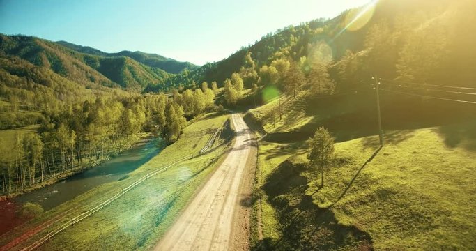 Aerial UHD 4K view. Low air flight over mountain rural dirt road and meadow at sunny summer morning. Near green trees, sun rays and mountain river