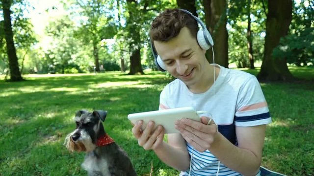 Boy watching something funny on tablet and sitting in the park with his dog, steadycam shot
