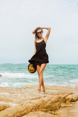 Beautiful fit lady in little black summer dress flying in the wind on big rocks touch her hairs. Beauty cute girl on a tropical beach sea ocean shore with large stones. Outdoor summer lifestyle.