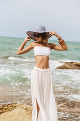 Sport lady stand on rock in white skirt and top, wearing sunglasses and correct her summer straw hat. Beauty cute girl on a tropical beach sea ocean shore with large stones. Outdoor summer lifestyle.