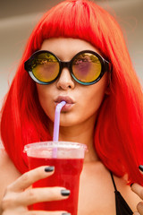 Close up portrait of a beautiful thirsty woman in red wig, black sexy swimsuit and fashion sunglasses drink red soda water with straw. Beauty cute girl on a tropical resort. Outdoor summer lifestyle.