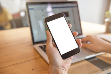 Close up of woman hand holding smart phone and using laptop at coffee shop, mobile with blank copy space white screen.