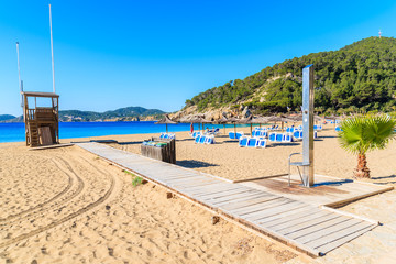Path to sandy beach with umbrellas and sunbeds in Cala San Vicente bay on sunny summer day, Ibiza...