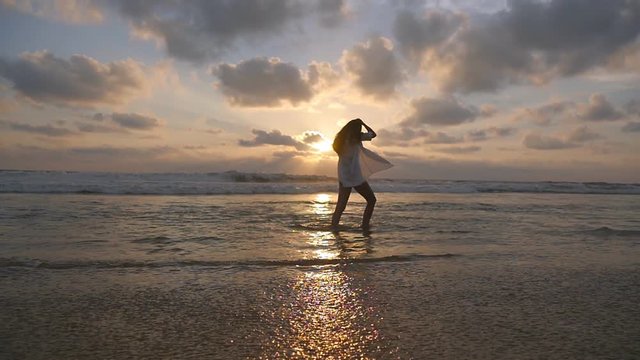 Happy woman in bikini and shirt walking at the ocean water on the beach at sunset. Young beautiful girl enjoying vacation and having fun at sea shore at sunrise. Summer holiday. Slow motion