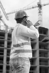 Black and white rear view image of construction engineer talking by phone and pointing at building site